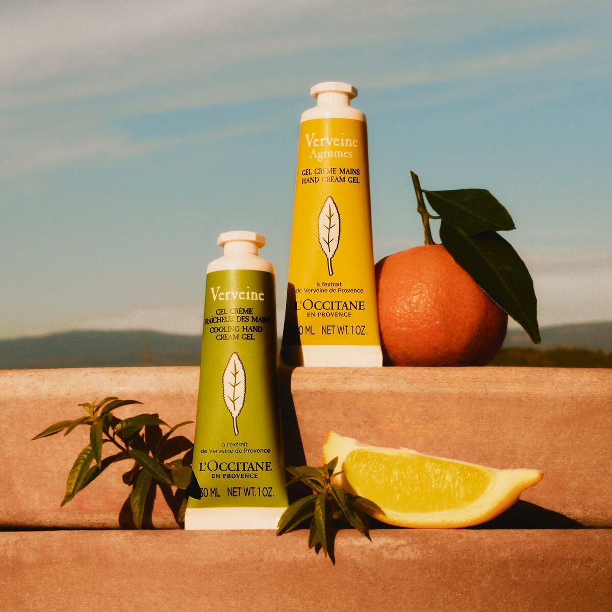 Two L'Occitane products with an orange and lemon on a wooden surface with a mountain background