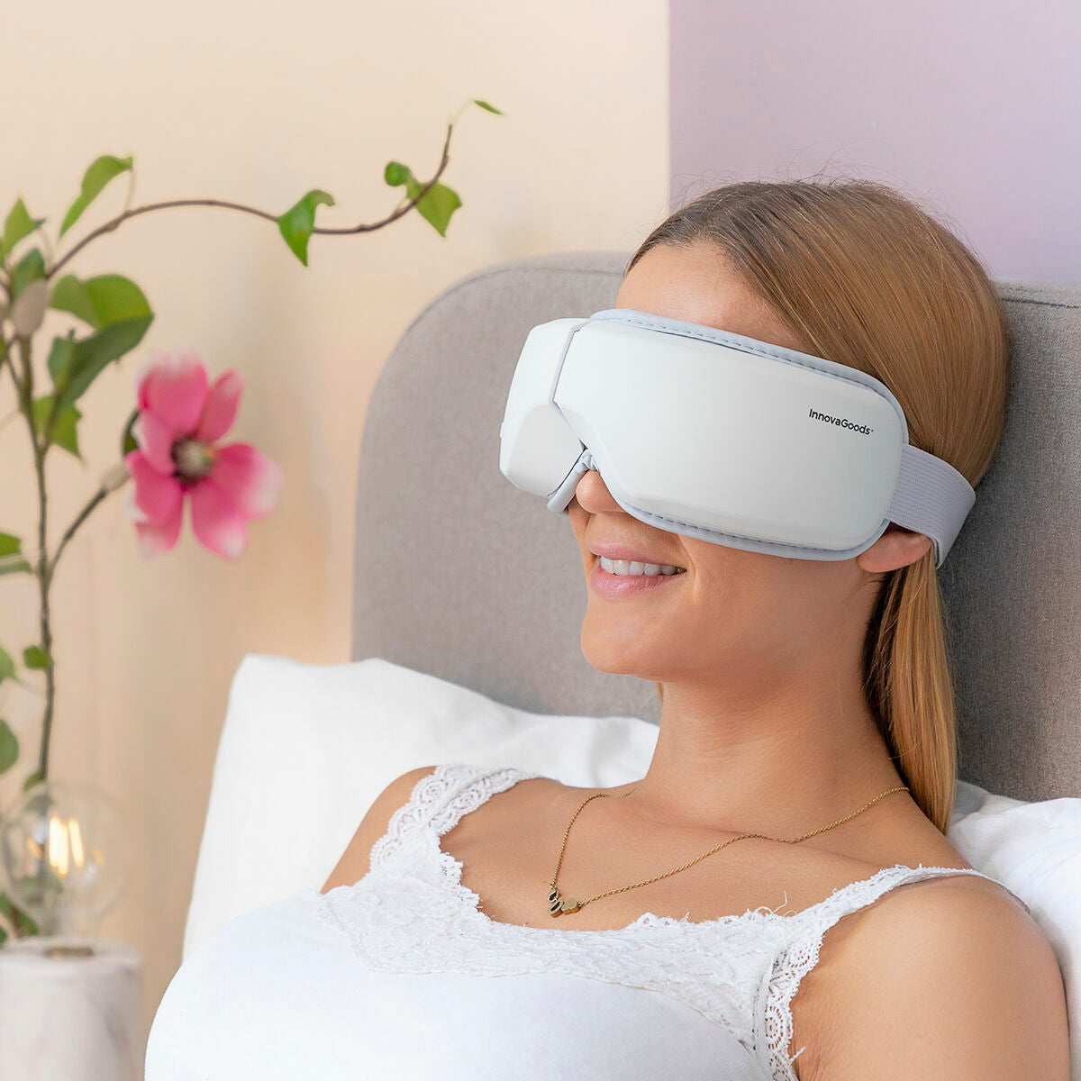 Woman wearing a white sleep mask in a bedroom setting with a plant and lamp in the background.